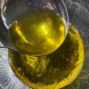 May include: A clear glass bowl and a stainless steel ladle filled with golden-yellow liquid. The ladle is pouring the liquid into the bowl. The bowl has a decorative, scalloped edge.