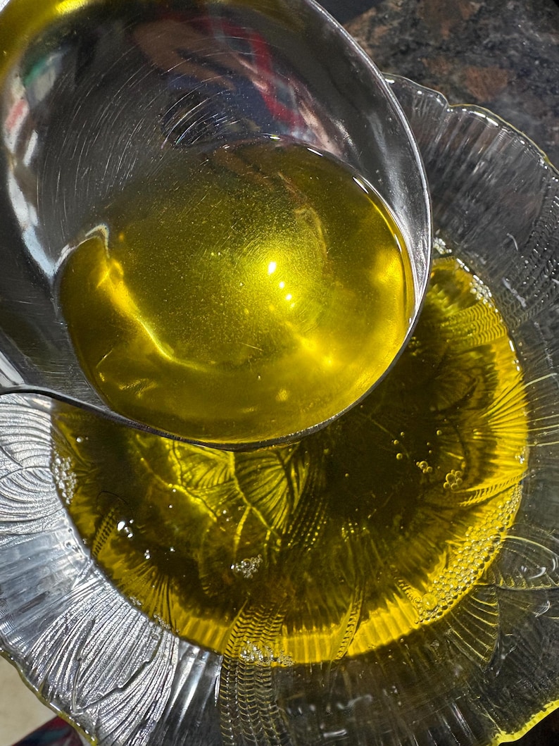 May include: A clear glass bowl and a stainless steel ladle filled with golden-yellow liquid. The ladle is pouring the liquid into the bowl. The bowl has a decorative, scalloped edge.