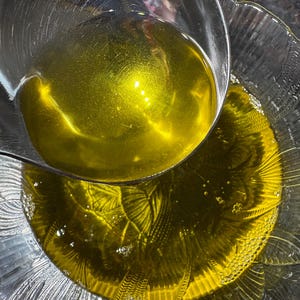 May include: A clear glass bowl is filled with golden-yellow liquid, possibly oil, with a metal ladle pouring more in. The bowl has a decorative, textured design. Bubbles are visible in the liquid. The image is a close-up shot.