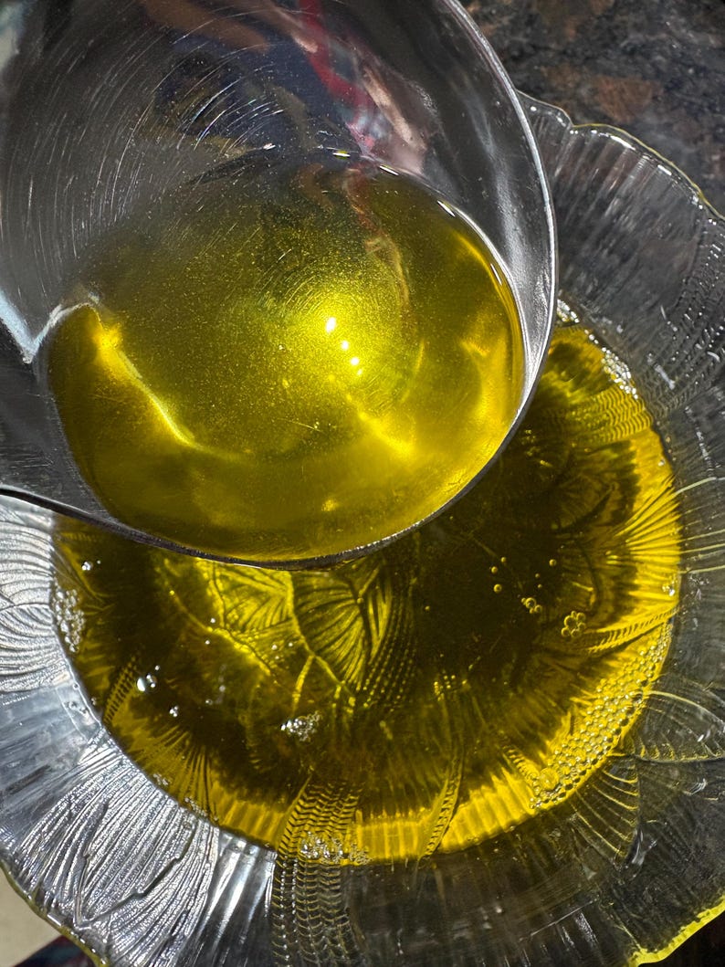 May include: A clear glass bowl is filled with golden-yellow liquid, possibly oil, with a metal ladle pouring more in. The bowl has a decorative, textured design. Bubbles are visible in the liquid. The image is a close-up shot.