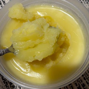 May include: A close-up of a container filled with a yellow, semi-solid substance, possibly ghee or clarified butter. A spoon scoops up a portion of the substance, revealing a textured, granular consistency. The container is clear, and the background is a patterned fabric.