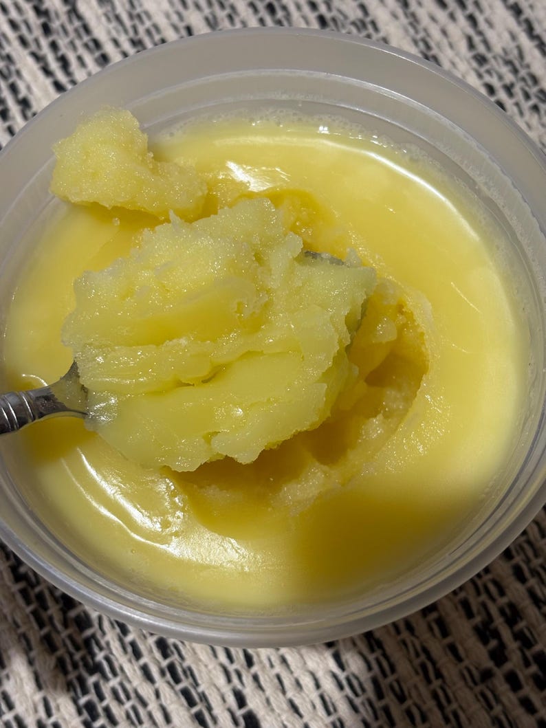 May include: A close-up of a container filled with a yellow, semi-solid substance, possibly ghee or clarified butter. A spoon scoops up a portion of the substance, revealing a textured, granular consistency. The container is clear, and the background is a patterned fabric.