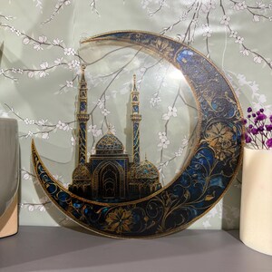 May include: A crescent moon-shaped wall decor with a gold and blue intricate design. The moon features a detailed silhouette of a mosque with minarets and domes.