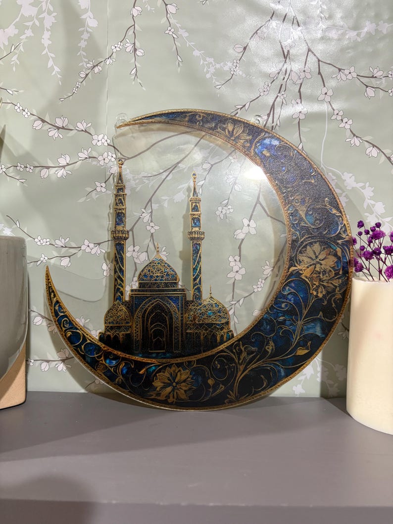 May include: A crescent moon-shaped wall decor with a gold and blue intricate design. The moon features a detailed silhouette of a mosque with minarets and domes.