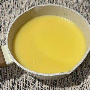May include: A small, light-coloured saucepan filled with a smooth, bright yellow liquid. The pan has a speckled, off-white exterior and a brown handle. The liquid fills the pan, suggesting cooking or food preparation.