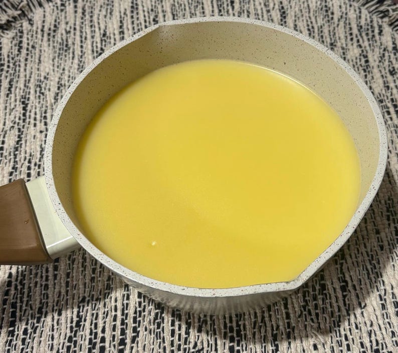 May include: A small, light-coloured saucepan filled with a smooth, bright yellow liquid. The pan has a speckled, off-white exterior and a brown handle. The liquid fills the pan, suggesting cooking or food preparation.
