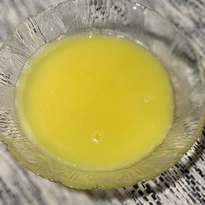 May include: A clear glass bowl containing a bright yellow liquid. The bowl has a textured rim. The liquid appears smooth and has a slight sheen, with a small bubble visible.