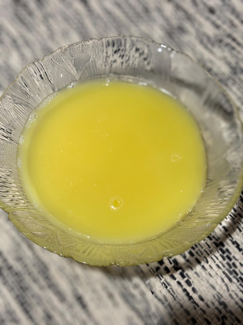 May include: A clear glass bowl containing a bright yellow liquid. The bowl has a textured rim. The liquid appears smooth and has a slight sheen, with a small bubble visible.