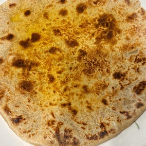 May include: Close-up of a round, golden-brown flatbread with darker, toasted patches. The bread has a slightly oily surface, indicating it's freshly cooked. The texture suggests a flaky or layered dough, possibly a paratha.