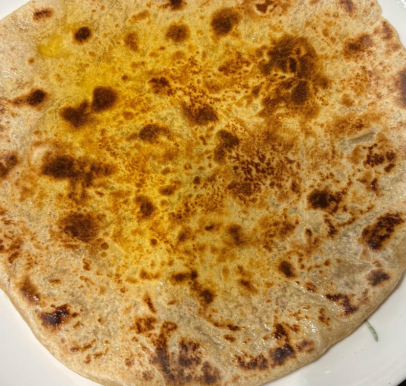 May include: Close-up of a round, golden-brown flatbread with darker, toasted patches. The bread has a slightly oily surface, indicating it's freshly cooked. The texture suggests a flaky or layered dough, possibly a paratha.