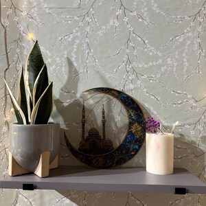 May include: A gray shelf with a potted snake plant, a decorative wooden crescent moon with a mosque design, and a white candle with purple flowers on top. The shelf is against a green wall with a white floral pattern.