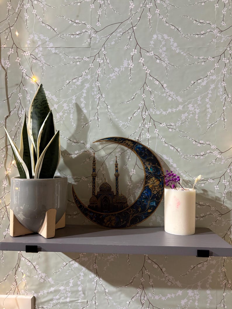 May include: A gray shelf with a potted snake plant, a decorative wooden crescent moon with a mosque design, and a white candle with purple flowers on top. The shelf is against a green wall with a white floral pattern.
