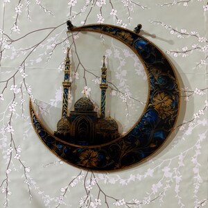May include: A decorative wall hanging featuring a crescent moon with intricate gold and blue designs. The moon is depicted with a mosque silhouette inside, symbolizing Islamic culture.
