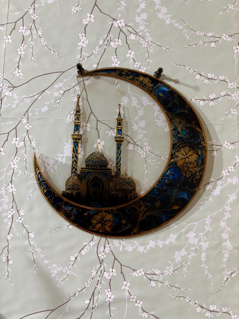 May include: A decorative wall hanging featuring a crescent moon with intricate gold and blue designs. The moon is depicted with a mosque silhouette inside, symbolizing Islamic culture.