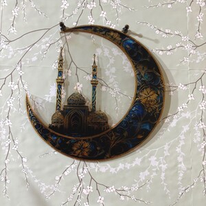 May include: A crescent moon-shaped wall decor with a gold and blue design featuring a mosque with minarets. The background is a light green with white floral branches.