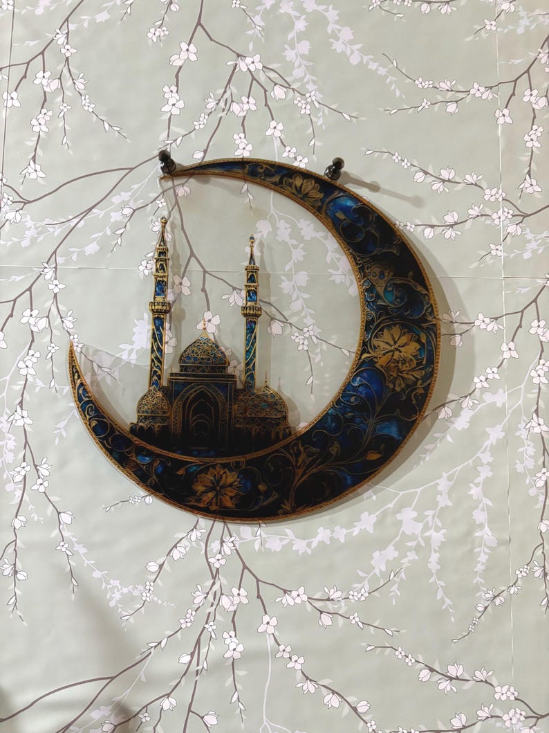 May include: A crescent moon-shaped wall decor with a gold and blue design featuring a mosque with minarets. The background is a light green with white floral branches.
