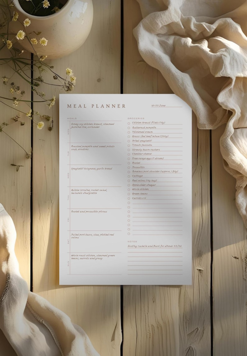 Image of a completed meal planner, sitting on a weathered wooden table. Nearby is dainty yellow flowers in a vase and a linen cloth. Light streams across the paper.