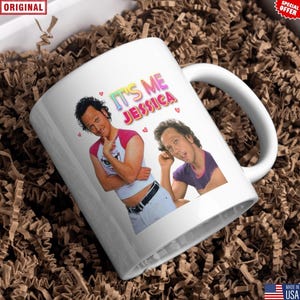 May include: White ceramic mug featuring a colorful graphic of a person with the text "IT'S ME JESSICA" and small pink hearts. The mug has a curved handle and is surrounded by brown packing material.