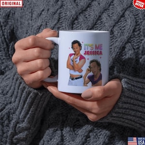 May include: White ceramic mug featuring two images of a man and the text "IT'S ME JESSICA" in pink and blue. The mug has a white handle and is held by a person wearing a gray sweater.