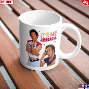 May include: White ceramic mug featuring a colorful design with the text "IT'S ME JESSICA" and images of a person. The mug has a white handle and is set on a wooden surface.