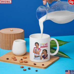 May include: A white ceramic mug with the text "IT'S ME JESSICA" and two images of a person. Milk is being poured into the mug from a glass pitcher. The mug is on a wooden tray with almonds and coffee beans.