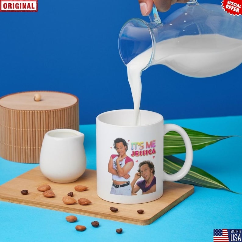 May include: A white ceramic mug with the text "IT'S ME JESSICA" and two images of a person. Milk is being poured into the mug from a glass pitcher. The mug is on a wooden tray with almonds and coffee beans.