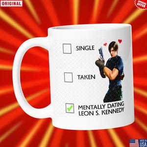 May include: White ceramic mug with a handle featuring a graphic of a man holding a gun, with the text "Single", "Taken", and "Mentally Dating Leon S. Kennedy". The mug has a red and yellow background.