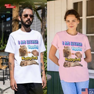 May include: White and pink t-shirts with the text "I AM EITHER Yapping Or Napping" and images of capybaras. The white shirt is worn by a person with a beard and sunglasses. The pink shirt is worn by a person with short hair.