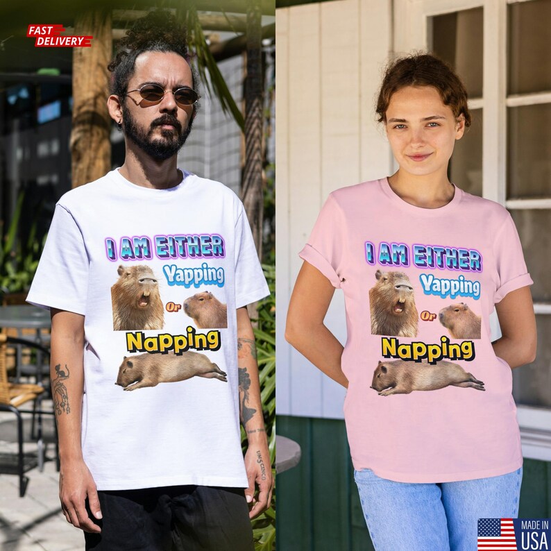 May include: White and pink t-shirts with the text "I AM EITHER Yapping Or Napping" and images of capybaras. The white shirt is worn by a person with a beard and sunglasses. The pink shirt is worn by a person with short hair.