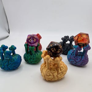 Op de afbeelding: Een verzameling kleurrijke dobbelsteenhouders, elk gevormd als een hersen met botachtige steunen. De dobbelstenen zijn in verschillende kleuren, waaronder paars, rood en oranje. De houders zijn geschilderd in tinten blauw, groen en goud.