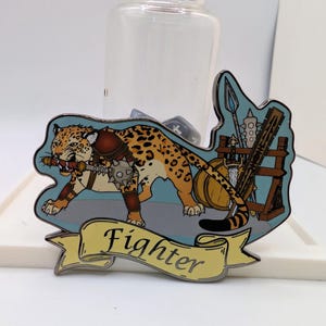 May include: A detailed enamel pin featuring a jaguar with orange and black spots, wearing armor and holding a weapon. The pin has a light blue border with various weapons and the word "Fighter" on a yellow banner.
