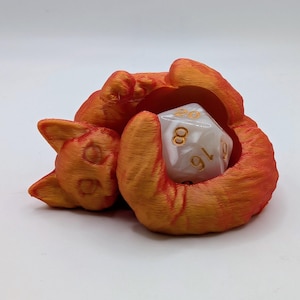 Op de afbeelding: Een oranje 3D-geprinte katvormige dobbelsteenhouder die een witte, veelzijdige dobbelsteen met gouden cijfers vasthoudt. De vorm van de kat toont afdruklijnen en de dobbelsteen wordt in de poten van de kat gehouden. Een speels en functioneel ontwerp.