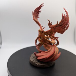 May include: A red and orange painted miniature figurine of a phoenix with outstretched wings. The phoenix is standing on a round base with a textured surface.