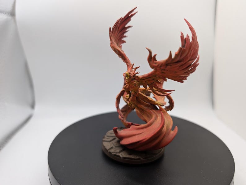 May include: A red and orange painted miniature figurine of a phoenix with outstretched wings. The phoenix is standing on a round base with a textured surface.