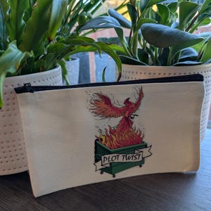 May include: A white canvas zippered pouch with a graphic of a phoenix rising from a green dumpster with the text "PLOT TWIST".