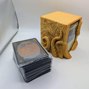 May include: A mustard yellow 3D-printed deck box featuring a textured surface with sculpted tentacles, holding a stack of trading cards. Beside it sits a separate, larger stack of Magic: The Gathering trading cards in clear protective sleeves.