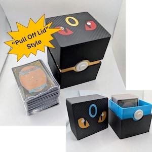 May include: A black square card storage box with a pull-off lid featuring red and yellow eye graphics and a central yellow ring. A stack of Magic The Gathering trading cards is shown next to the box, which includes a blue inner tray component.