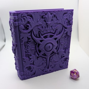 May include: A purple, ornate book-shaped box with a detailed relief design. The box features a central eye symbol and floral patterns. A purple and gold 20-sided die is next to the box. The box is likely for storing tabletop gaming accessories.