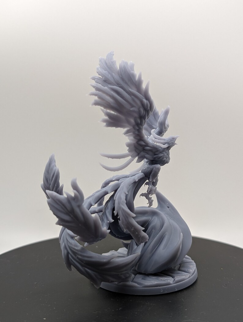 May include: A gray 3D printed miniature of a phoenix with its wings spread wide. The phoenix is perched on a base with a swirling design.