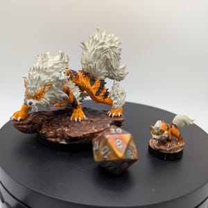 May include: Two painted figurines of a fire-type Pokemon, Arcanine, one larger than the other. The larger Arcanine is standing on a brown base with a 20-sided die in front of it. The smaller Arcanine is standing on a smaller brown base.