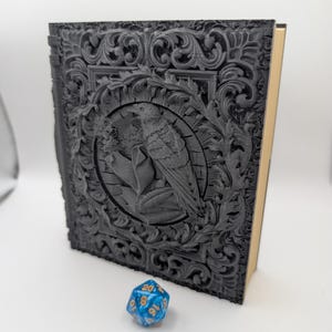May include: A dark grey, intricately carved book with a raven design in a circular frame. The cover features ornate floral and scrollwork. The book has gold-coloured page edges and a blue and white twenty-sided die is in the foreground.