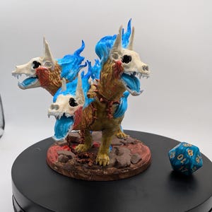 Peut inclure: Une figurine de chien à trois têtes avec des flammes bleues et une chaîne autour du cou. La figurine est peinte dans des tons de brun, blanc et bleu. La figurine est posée sur une base brune avec des accents rouges. Un dé bleu et or à vingt faces est au premier plan.