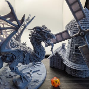 May include: A detailed gray dragon figurine with large, textured wings stands beside a miniature windmill. An orange and yellow twenty-sided die rests in the foreground. The scene evokes fantasy and tabletop gaming themes.