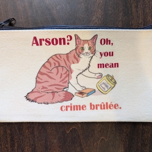 May include: A white canvas zippered pouch with a red tabby cat illustration. The cat is holding a box of matches and a jar of candle wax. The text on the pouch reads "Arson? Oh, you mean crime brûlée."