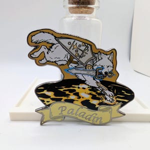 May include: A detailed enamel pin featuring a white wolf in action, wielding a sword and shield. The wolf is set against a gold and black background, with the word "Paladin" on a banner below. The pin is a collectible item.