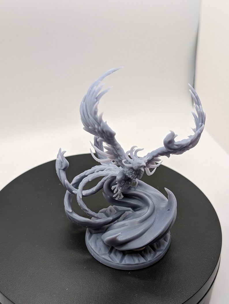 May include: A gray 3D printed miniature of a phoenix with its wings spread wide. The phoenix is standing on a circular base with a swirling design.