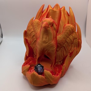 May include: A 3D-printed phoenix-shaped dice holder in shades of orange and gold. The bird has outstretched wings and is surrounded by flame-like structures. A multi-sided die rests in the base of the holder.