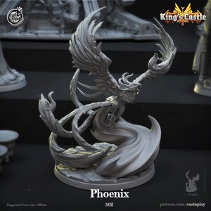 May include: A gray 3D printed miniature of a phoenix in flight. The phoenix has detailed wings, a long tail, and a fierce expression. The miniature is on a round base with a textured surface. The text "Phoenix 502" is printed below the miniature.