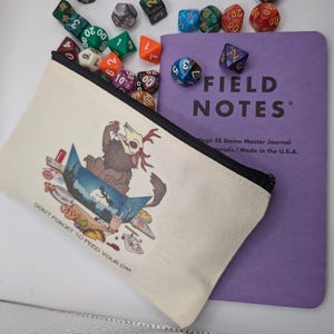 Könnte beinhalten: Eine weiße Stofftasche mit einem schwarzen Reißverschluss und einer Cartoon-Illustration eines Wesens mit Geweih. Die Tasche ist mit "Don't forget to feed your DM" beschriftet. Die Tasche liegt neben einem violetten Notizbuch mit dem Text "FIELD NOTES" und "Page SE Game Master Journal" auf dem Cover.