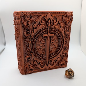 May include: A brown, 3D-printed book-shaped container with intricate, embossed designs and a sword emblem. A twenty-sided die rests beside the container. The container is likely for storing items or as a decorative piece.